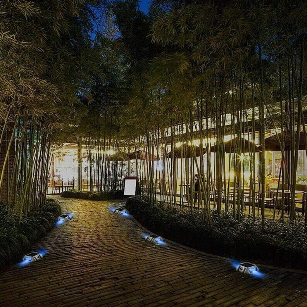 Brick-paved pathway lined with blue solar-powered driveway deck marker lights—illuminating a scenic route bordered by tall bamboo and leading to a cozy, well-lit building with large glass windows and outdoor seating, enhancing safety and ambiance during nighttime.