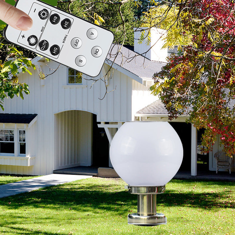Solar-powered white globe garden light mounted on a metal base in front of a house, with a hand holding a remote control showcasing brightness, timer, and color mode settings.