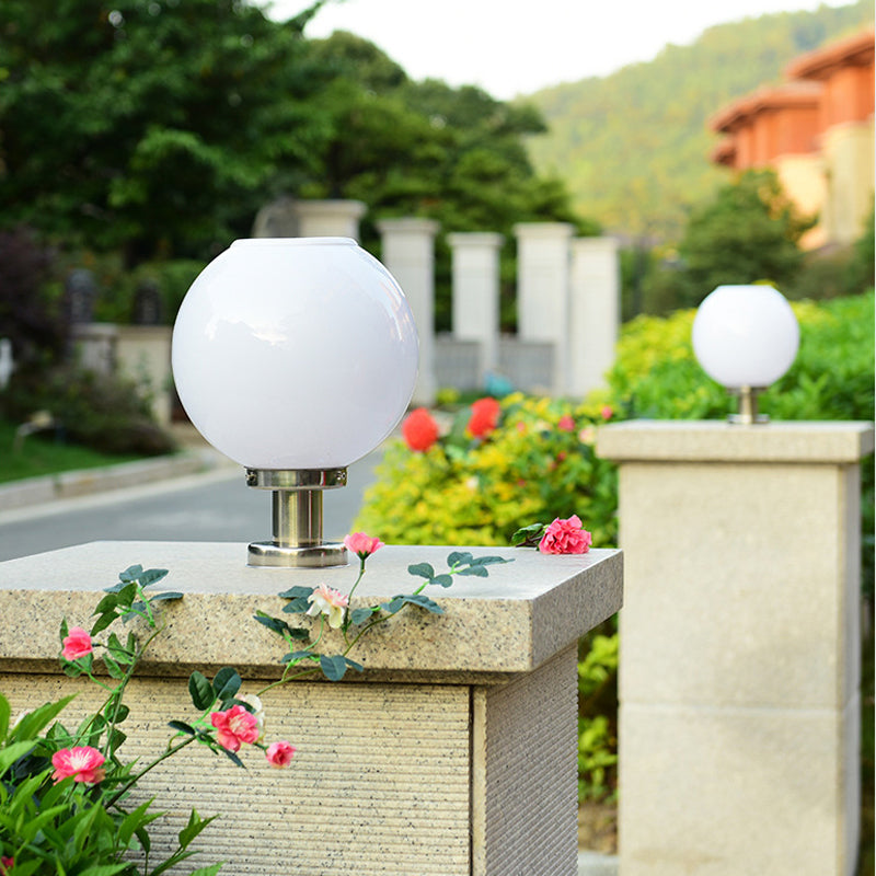 White solar-powered spherical globe light with stainless steel base, mounted on a stone pillar in an outdoor garden setting, surrounded by lush greenery and blooming flowers