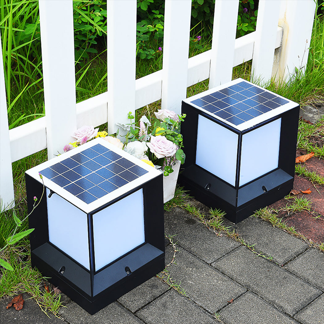 Two square solar pillar lights with frosted white side panels and sleek black metal frames, positioned beside a white picket fence and decorative plants. Each light features a top-mounted solar panel and is designed for outdoor use—ideal for garden paths, gates, or patio lighting with dusk-to-dawn activation and remote-controlled triple-color LED options.