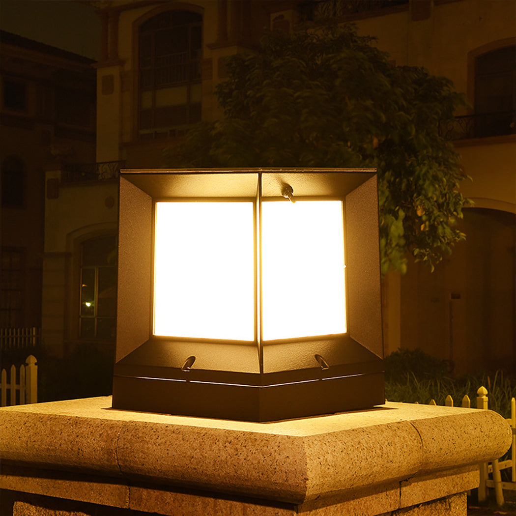 Modern square solar pillar light mounted on a stone pedestal, casting a warm golden glow across a landscaped courtyard. The fixture features a top solar panel and minimalist black frame, ideal for outdoor garden or driveway installations where ambient elegance meets energy-efficient design. The light is currently set to warm white, enhancing the cozy atmosphere of the space.