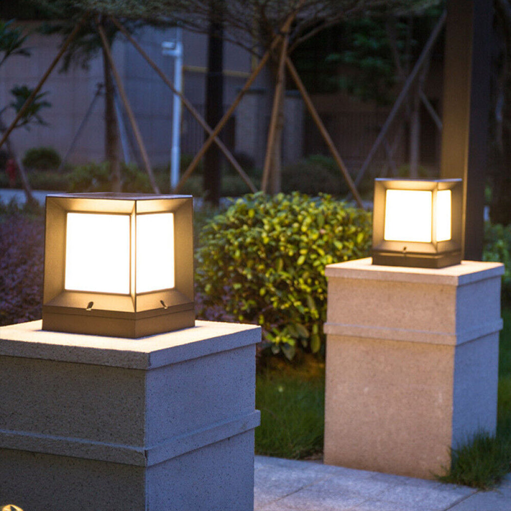 Two modern square solar pillar lights with warm white illumination, positioned on stone columns within a lush landscaped garden. Featuring sleek black frames and top-mounted solar panels, the lights cast a welcoming glow onto surrounding greenery and trees—perfect for enhancing ambiance along pathways, driveways, or courtyard entrances.