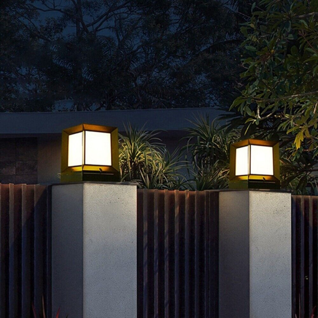 Two square solar pillar lights mounted on concrete fence columns, glowing in warm white mode against a nighttime garden backdrop. Each light features a sleek black housing with a top solar panel, providing ambient illumination for outdoor spaces such as garden entrances, pathways, or perimeter fencing.