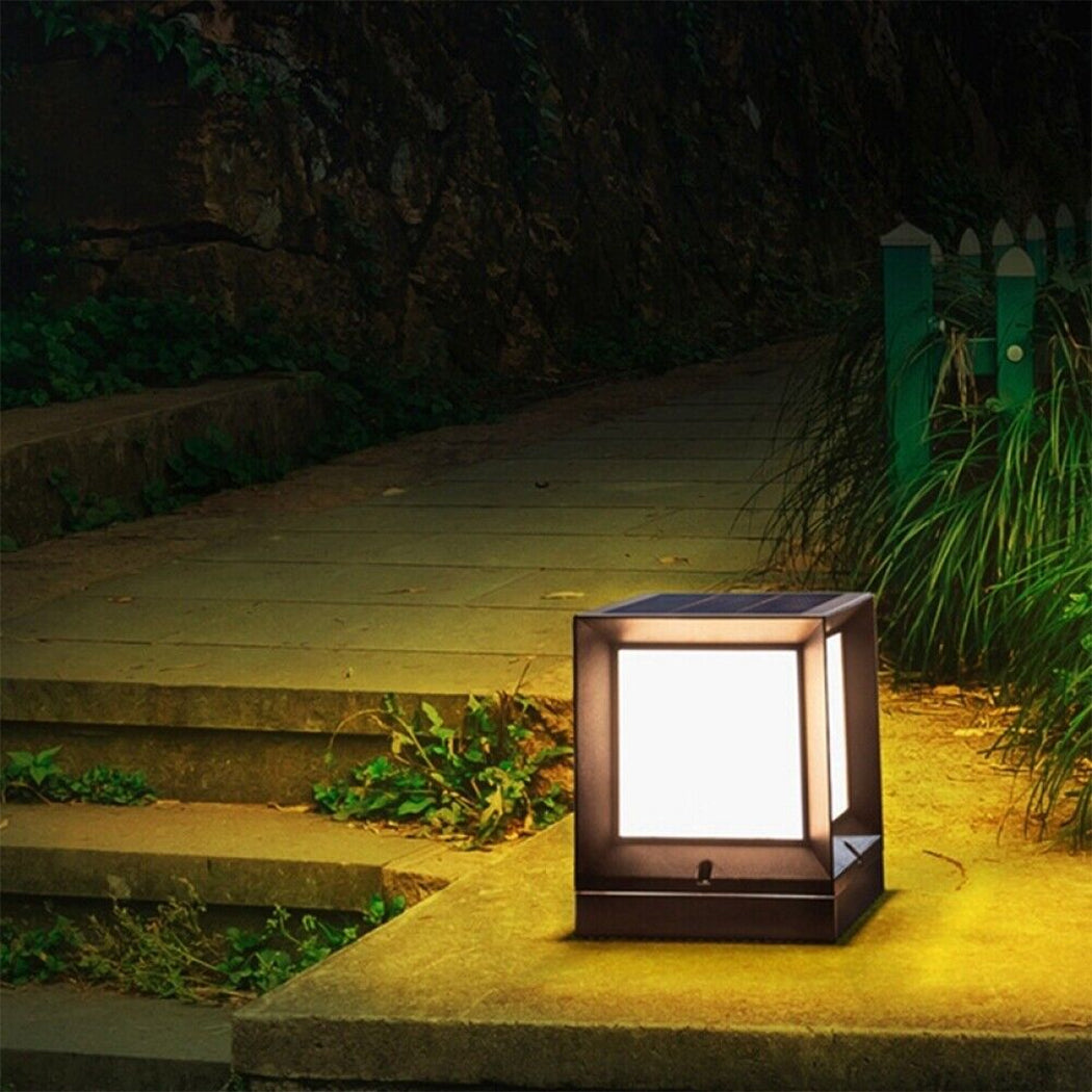 Square solar pillar light with sleek black housing illuminated in warm white mode, mounted on a stone pathway bordered by greenery and low fencing. The fixture provides focused lighting in a dim outdoor setting, highlighting its effectiveness for nighttime visibility along steps or garden borders. Top-mounted solar panel ensures eco-friendly operation, ideal for residential landscaping.