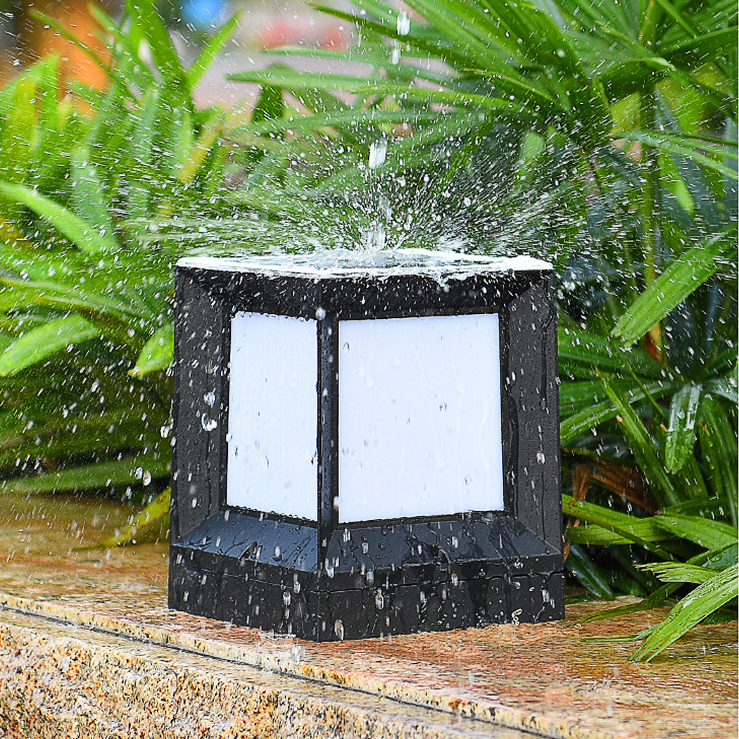 Square solar pillar light with black frame and white side panels, shown mid-splash as water cascades off its top and sides—demonstrating waterproof performance. Positioned on a stone surface with lush green foliage in the background, this fixture combines durability with sleek design, ideal for outdoor garden, fence, or patio installations exposed to rain.