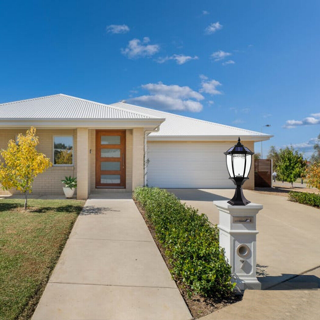 Antique-style solar post lamp with ornate detailing and a warm glow, mounted on a stone pillar at the edge of a garden path, surrounded by greenery and a modern home in the background