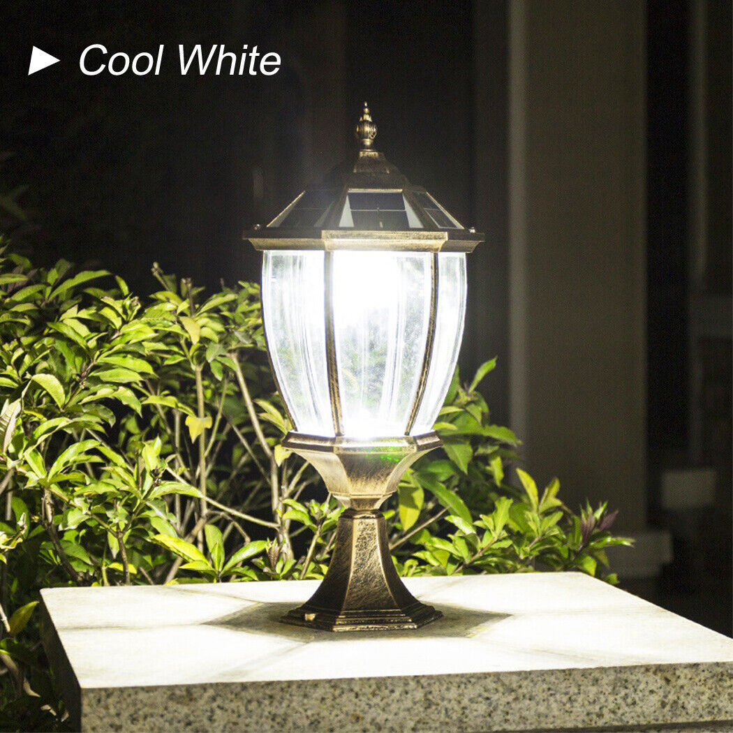 Classic bronze solar post lamp with clear glass panels, glowing with cool white light, mounted on a stone base and surrounded by lush green foliage in a garden setting.