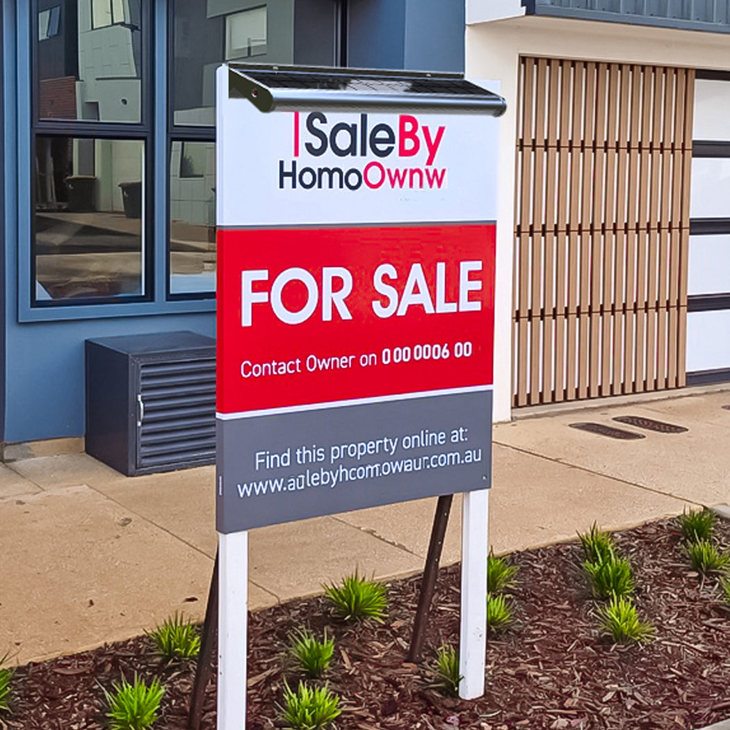 "For sale billboard installed in front of a modern residential property, featuring a compact solar-powered LED light mounted above the sign. The light includes a small solar panel and weatherproof casing, positioned to illuminate the billboard during nighttime. The setup highlights energy-efficient outdoor signage lighting