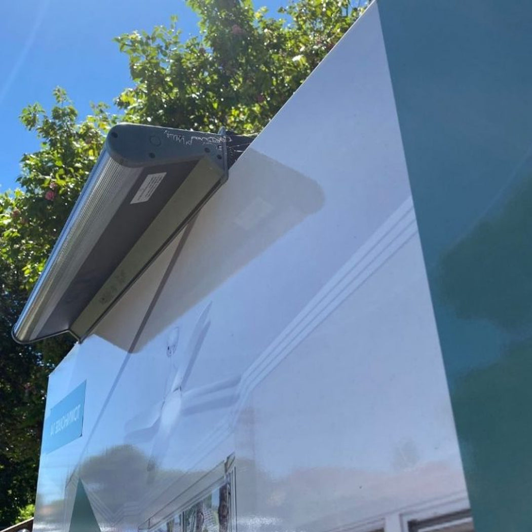 Outdoor real estate billboard equipped with a solar-powered LED light mounted at the top edge, angled downward to illuminate the sign. The compact fixture features a built-in solar panel and weather-resistant casing, providing energy-efficient nighttime visibility for property advertisements. Set against a clear sky and greenery, the setup highlights sustainable lighting for outdoor signage