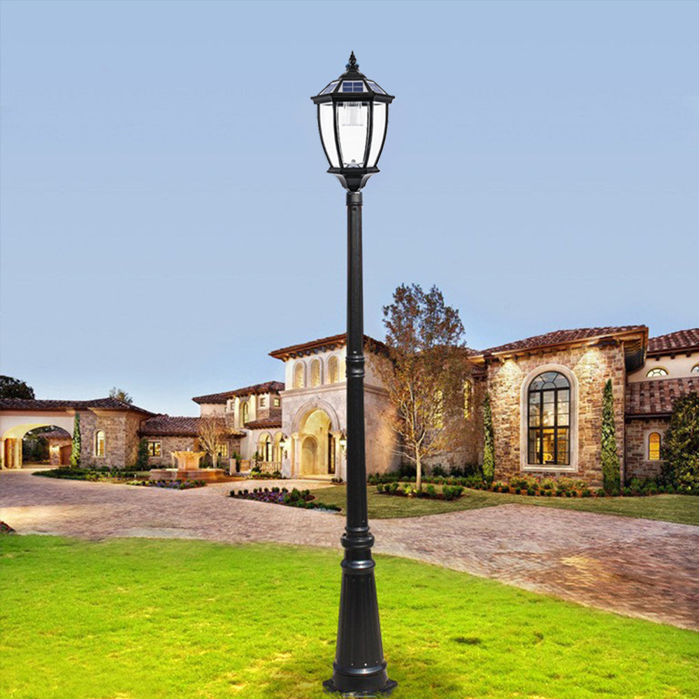 A classic black streetlight with a lantern-style top stands illuminated in a picturesque residential area. In the background, a large, elegant house featuring stone walls, arched windows, and a tiled roof is surrounded by a neatly landscaped garden with green grass and trees. The clear blue sky adds to the serene and charming atmosphere.