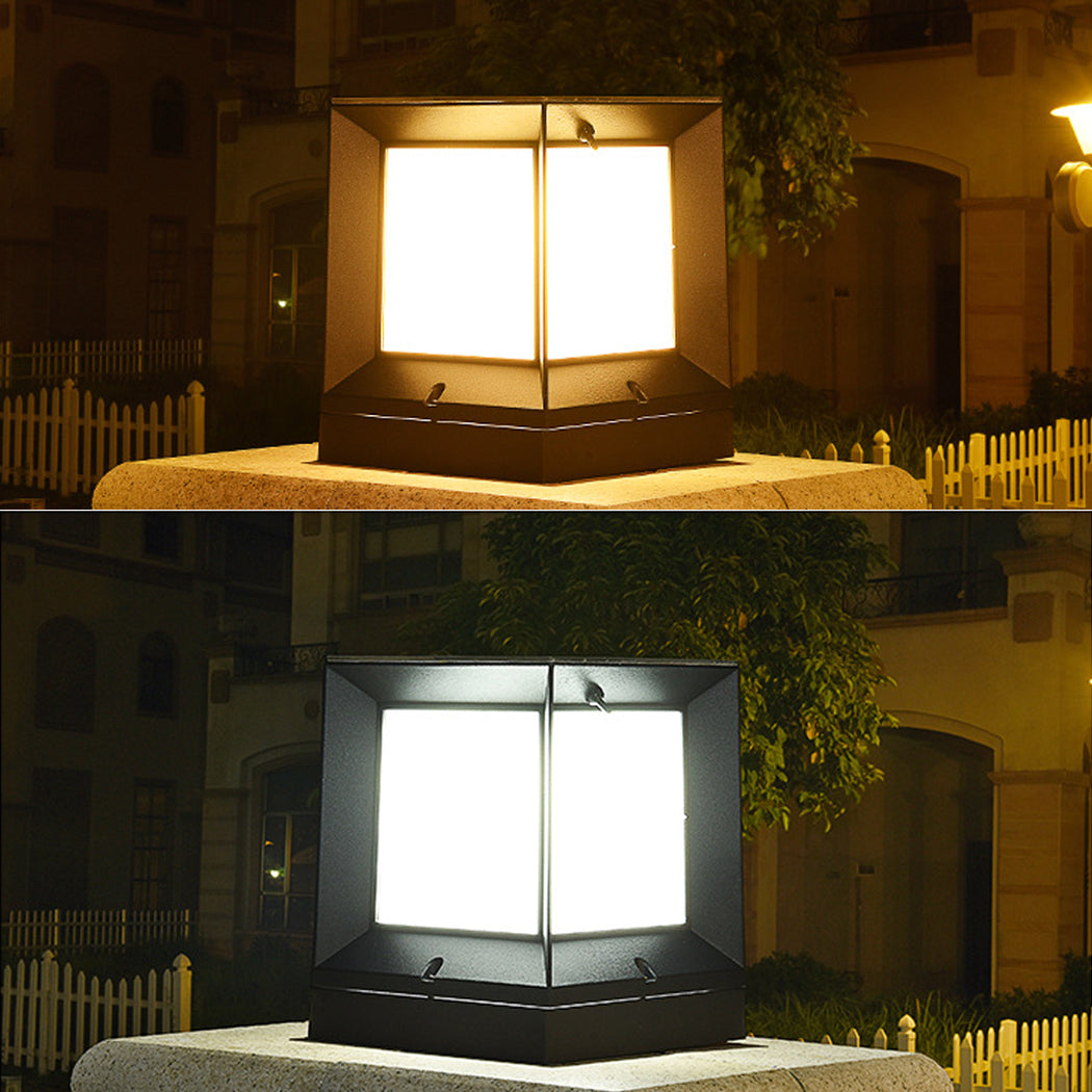 Two square solar pillar lights displayed side-by-side atop a single stone column, showcasing their dual lighting modes—warm yellow and cool white. Each fixture features a sleek black frame with a top-mounted solar panel and minimalist geometric design, set against a background of picket fencing, greenery, and a building exterior. Ideal for highlighting color temperature options and comparing ambient effects for garden or gate lighting installations.