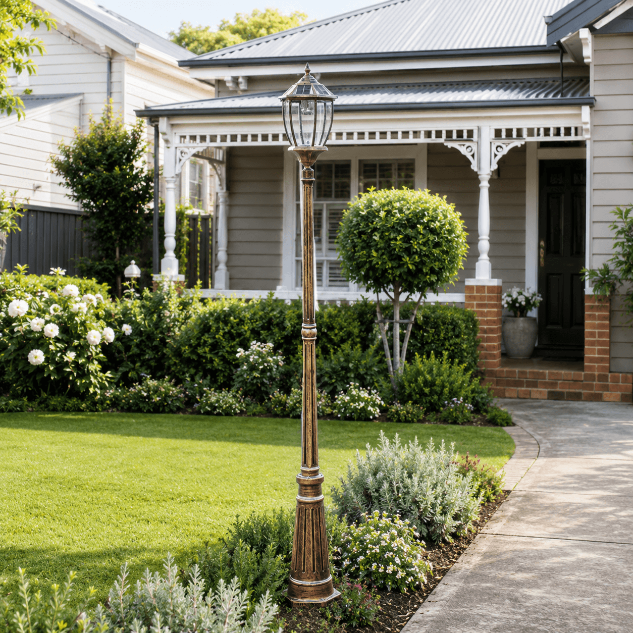 A bronze solar driveway post light standing in a front yard garden bed next to a paved concrete walkway entry.