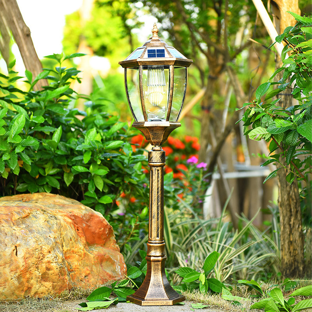 Decorative bronze solar post light with an antique clear glass lantern illuminated in a lush green garden setting next to landscaping rocks.