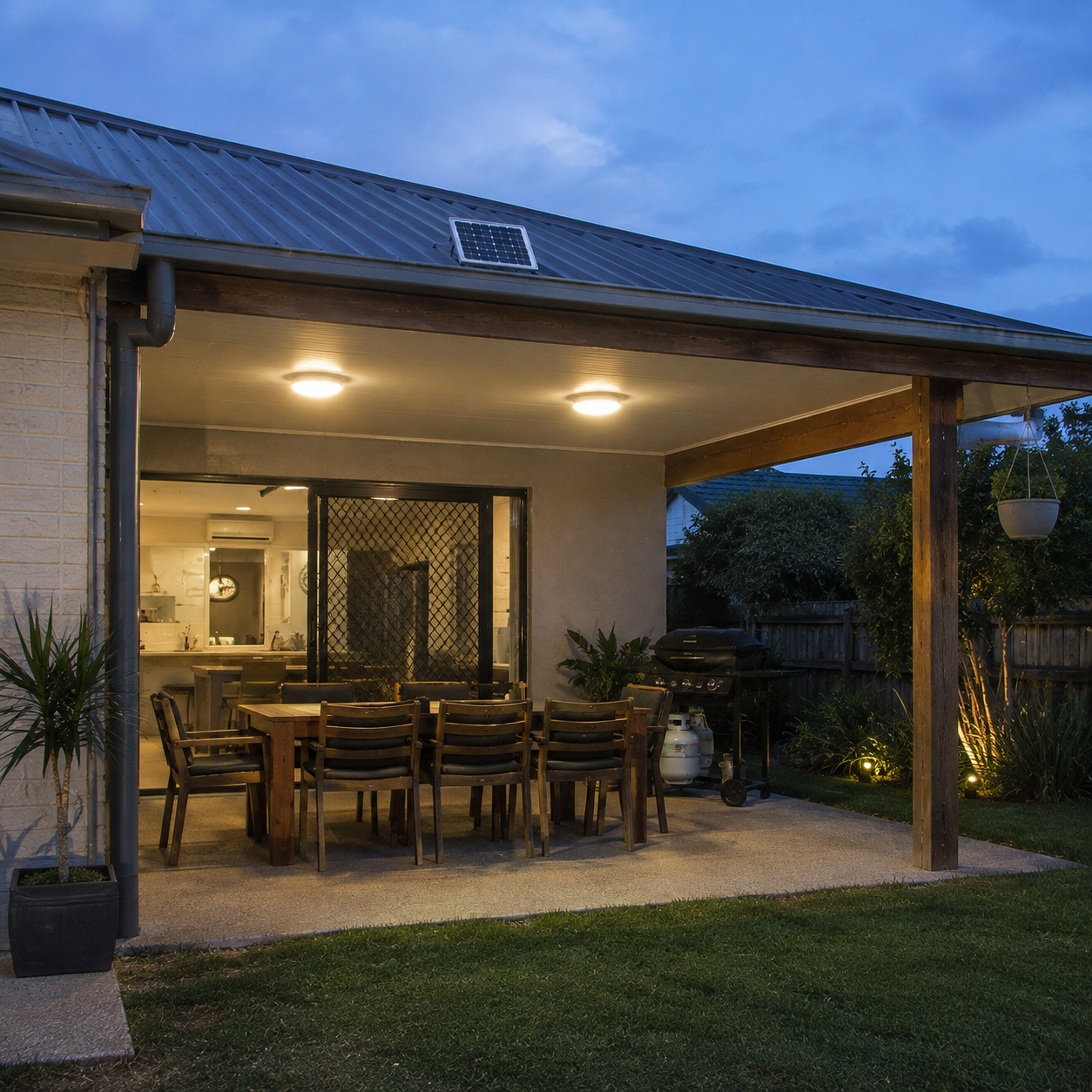 An Australian outdoor backyard patio veranda lit up at dusk by a dual solar ceiling light system with a small roof panel and an outdoor timber dining table set up for entertainment.