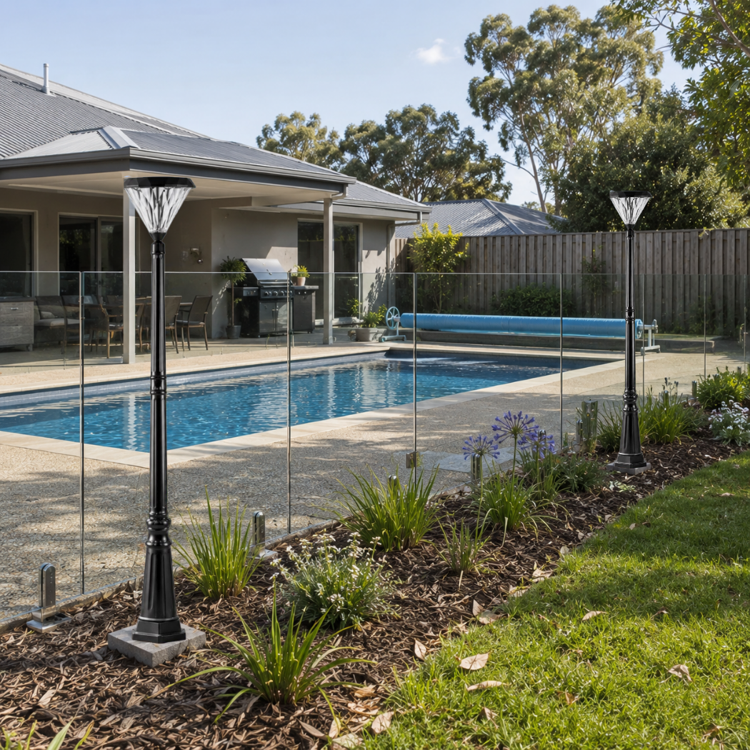 Two modern flat top solar lantern poles installed along a glass pool fencing perimeter and landscaped garden bed near an outdoor alfresco patio area.