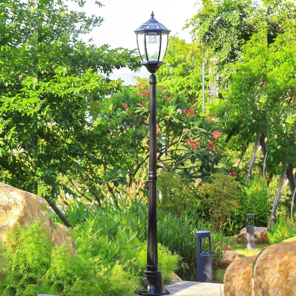 Decorative solar garden lamp post with an antique clear glass lantern standing surrounded by green trees and landscape garden rocks.