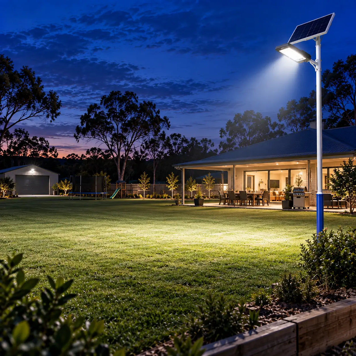 A Rainman G60 commercial solar street light pole-mounted next to a large residential homestead backyard lawn, brightly illuminating an outdoor veranda entertainment area and backyard garden at night.