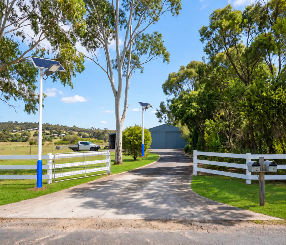 Two Rainman G60 commercial solar street lights pole-mounted along a concrete driveway entrance of an Australian acreage property, providing standalone lighting near a white post-and-rail fence, rural farm gate, and a large machinery shed.