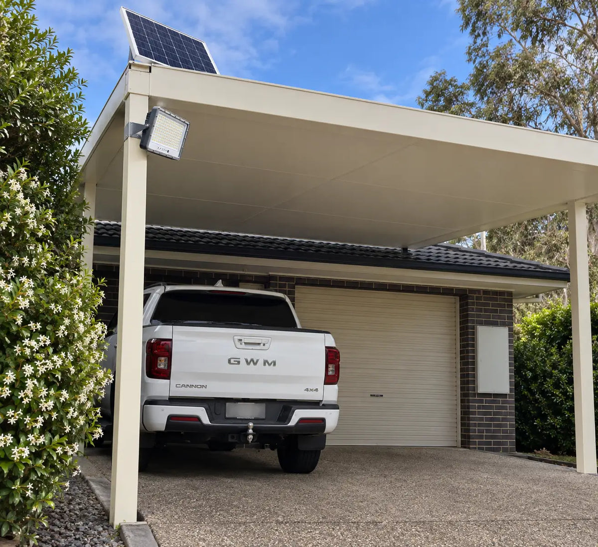 A Rainman K30 residential solar flood light and compact monocrystalline solar panel mounted onto a modern cream-coloured flat roof carport beam, providing off-grid security lighting over a concrete garage driveway and parked 4x4 utility vehicle during the day.