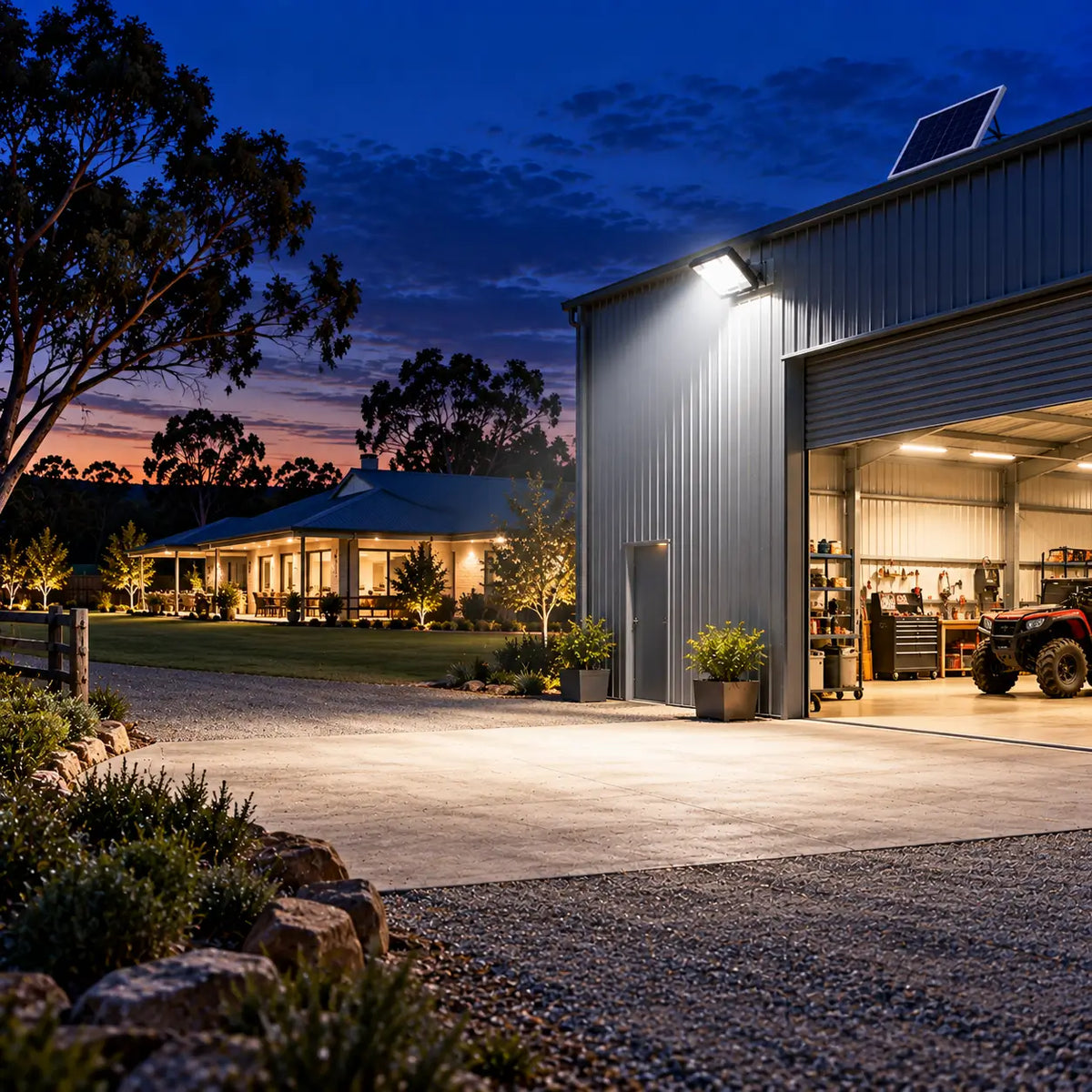 A Rainman K90 commercial solar flood light wall-mounted on a metal farm shed casting a bright wide angle security beam over a concrete apron driveway with a rural acreage homestead house visible in the background at sunset.