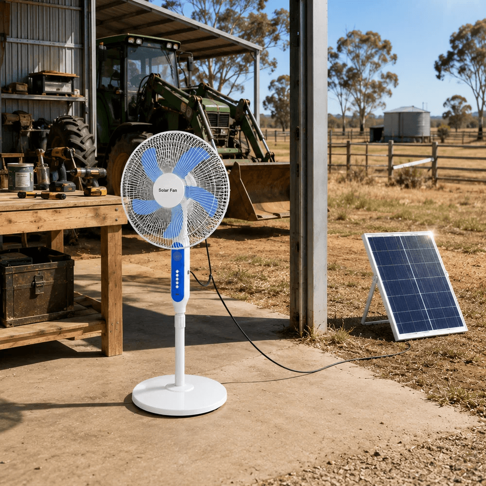 A white and blue solar pedestal fan positioned inside an Australian farm machinery shed next to a workbench, with its solar panel setup charging on the ground outside.