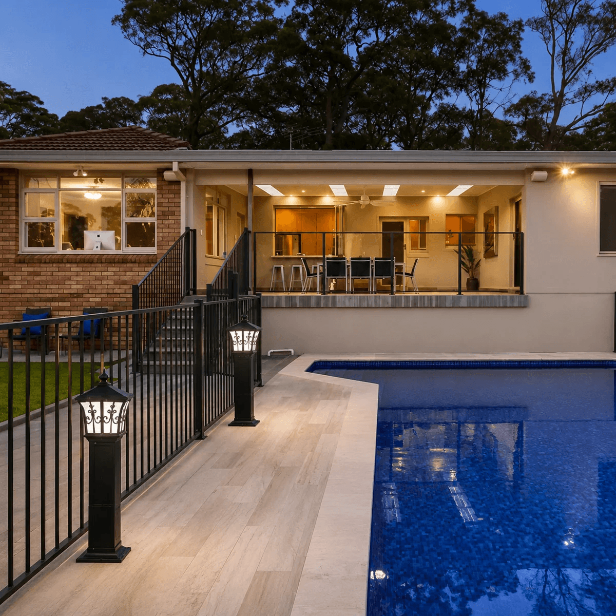 Solar pillar light auto on at dusk feature illuminating a concrete poolside patio and backyard entertaining area under warm white LED glow at night.