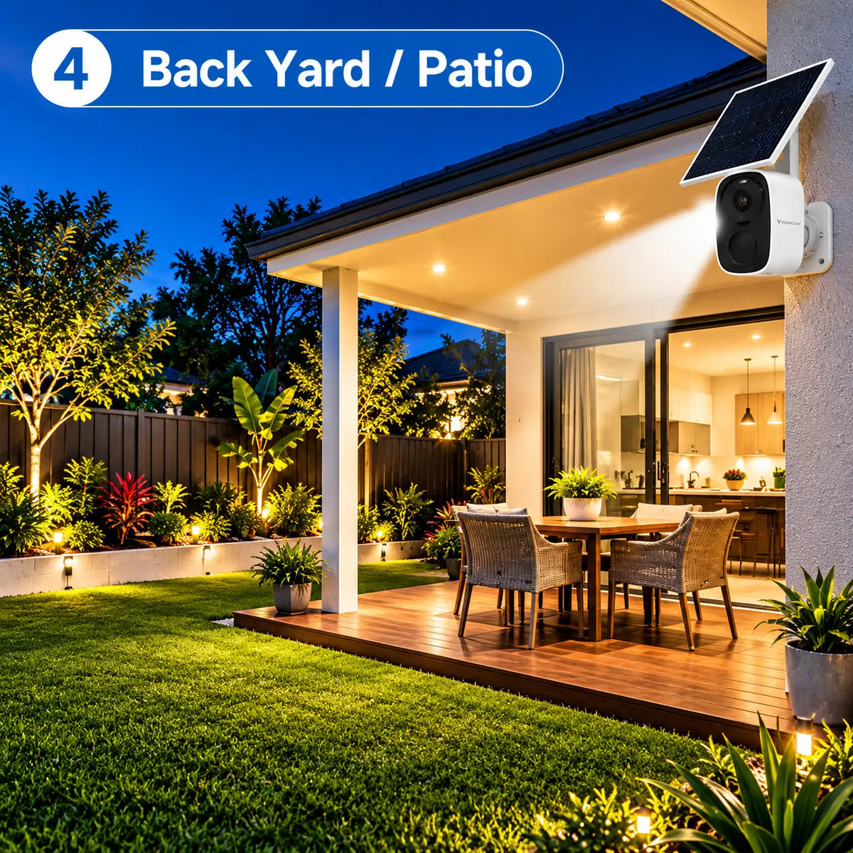 A Vstarcam BG55 solar powered security camera wall-mounted over a luxury timber deck patio, casting a bright security spotlight beam down onto an outdoor dining table, lawn grass backyard, and perimeter fence at night.