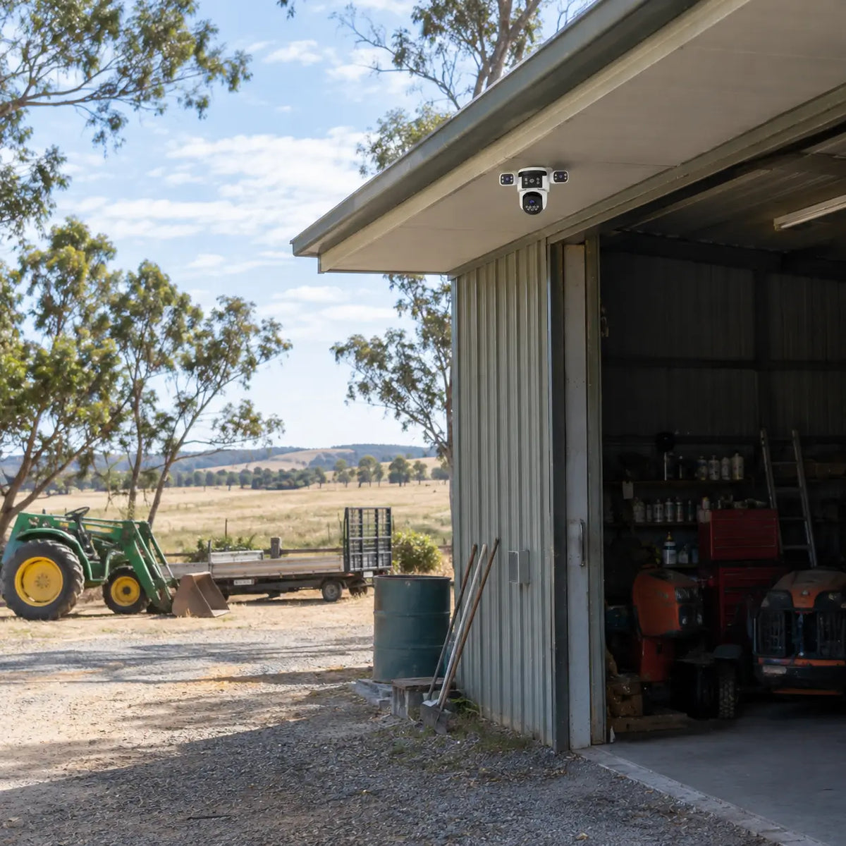 A real-world daytime installation of the VStarcam CS621SR three-lens security camera mounted under a corrugated steel farm machinery shed eave, monitoring an open workshop storage bay, parked tractor, and rural acreage paddock.