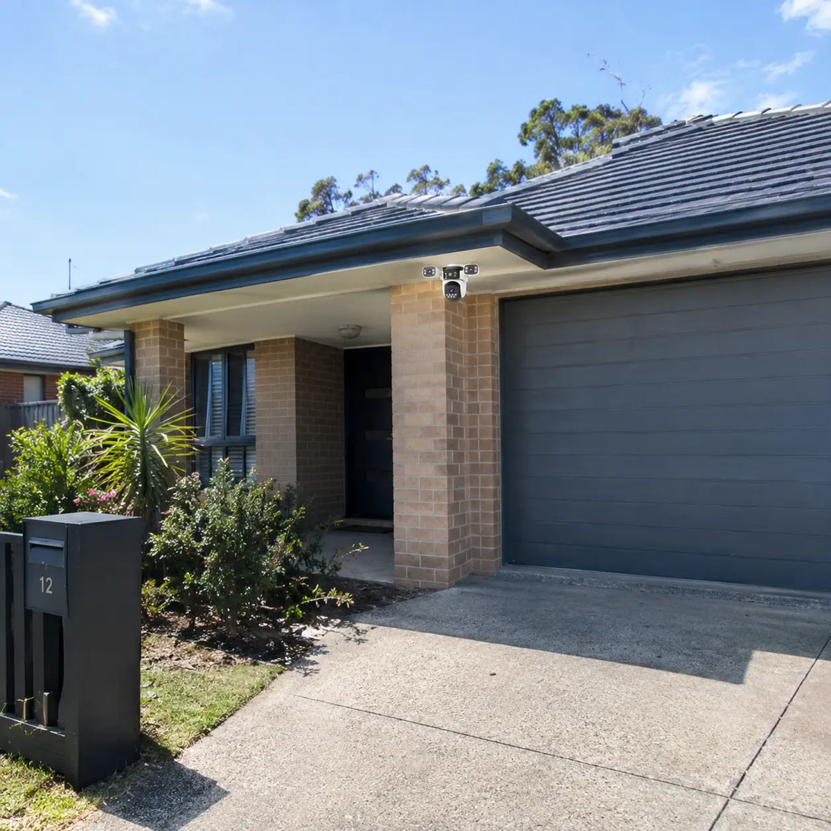 A real-world daytime installation of the VStarcam CS621SR three-lens security camera mounted on a brick support pillar, monitoring a concrete driveway apron and a charcoal sectional garage door.