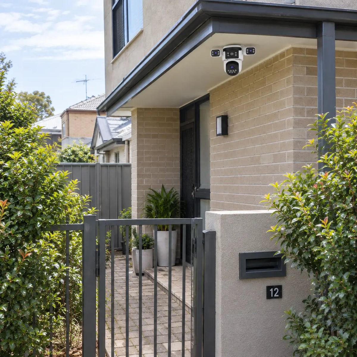 A real-world daytime installation of the VStarcam CS621SR three-lens security camera mounted under a two-story home awning, monitoring a charcoal metal entry gate, render boundary pillar with letterbox, and front courtyard path.